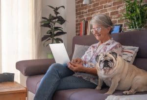 Smiling elderly woman at home working with laptop, sitting close to her old pug dog going online to get her Medicare and You Book